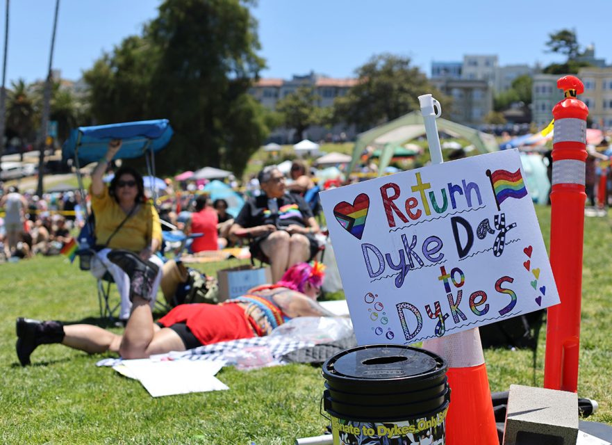 People sitting and lying on the grass at an outdoor event; a sign reading "Return Dyke Day to Dykes" with heart and rainbow decorations is prominently displayed in the foreground.