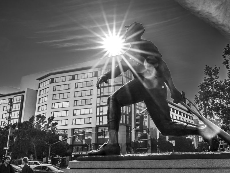 A black and white photo of a large statue of a person holding a bat, with the sun shining behind it. Buildings and pedestrians are visible in the background.