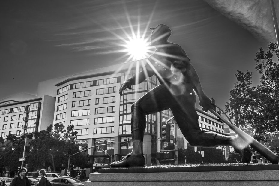A black and white photo of a large statue of a person holding a bat, with the sun shining behind it. Buildings and pedestrians are visible in the background.