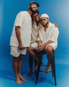 Three men standing in front of a blue background, with one sitting on a stool. Two men are smiling while one looks serious. All three are dressed in light-colored clothes.