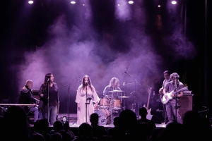 A band is performing on a dimly lit stage with purple spotlights. There are six musicians, including a drummer, keyboardist, guitarist, and singers. The audience is visible in the foreground.
