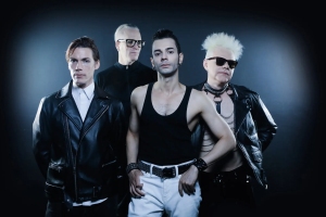 Four men stand together against a dark background. Three wear black leather jackets, and one in the center wears a black tank top and white pants. One man has spiked white hair and sunglasses.