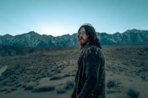 A person with long hair and glasses stands in a vast, barren landscape with mountains in the distance.