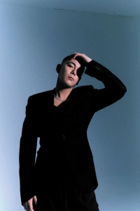 Person with short hair in a black suit poses against a light blue background, resting their hand on their head.