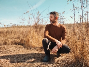 A man wearing a brown sweater and black pants is squatting in a dry, grassy field under a clear blue sky.