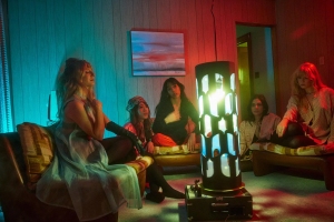 Five women sitting on couches in a dimly lit room with a cylindrical light fixture in the center. The room has colored lighting and a painting on the wall.