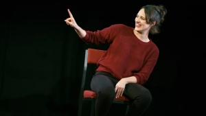 A person sitting on a red chair, pointing with their index finger, and smiling against a dark background. They are wearing a red sweater and black pants.