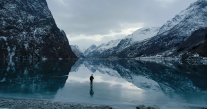 A lone figure stands on a frozen lake surrounded by snow-covered mountains under a cloudy sky.