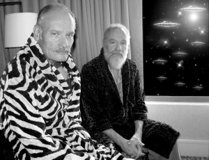 Two men in robes sit indoors by a curtain-covered window showing a night sky with numerous flying saucers outside.