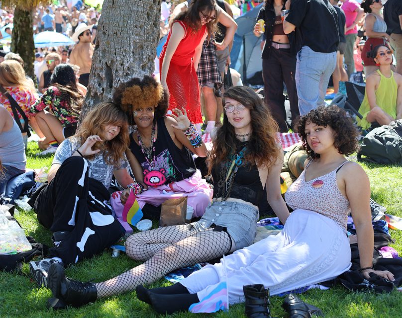 Four people sitting on the grass at an outdoor event, with one holding a small flag. They are surrounded by other attendees.