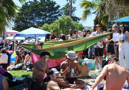 A group of people relax in a park. Some sit or lounge on the grass and others stand around a green and yellow hammock. Trees and a blue sky are visible in the background.