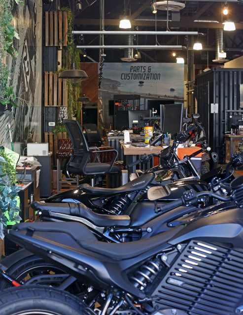 An indoor motorcycle shop with several black Harley Davidson motorcycles lined up in the foreground and a workbench with tools, plants, and a sign that reads "Parts & Customization" in the background.