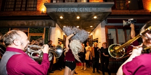 A band performs with brass instruments while a dancer engages the crowd outside Foreign Cinema, a venue with a marquee. People are gathered and watching.