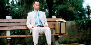 A man in a white suit and blue plaid shirt sits on a wooden bench outdoors with a suitcase and a box of chocolates beside him.