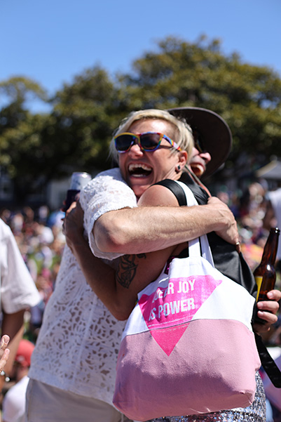 Two individuals embrace outdoors during a sunny day, both smiling. One holds a bag with a pink triangle and text that reads, "Your joy is power." A bottle is visible in the other person's hand.