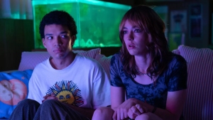 Two people sit closely on a couch in a dimly lit room with a colorful fish tank behind them. The person on the left is wearing a white t-shirt with a sun graphic, and the person on the right is wearing a dark shirt.