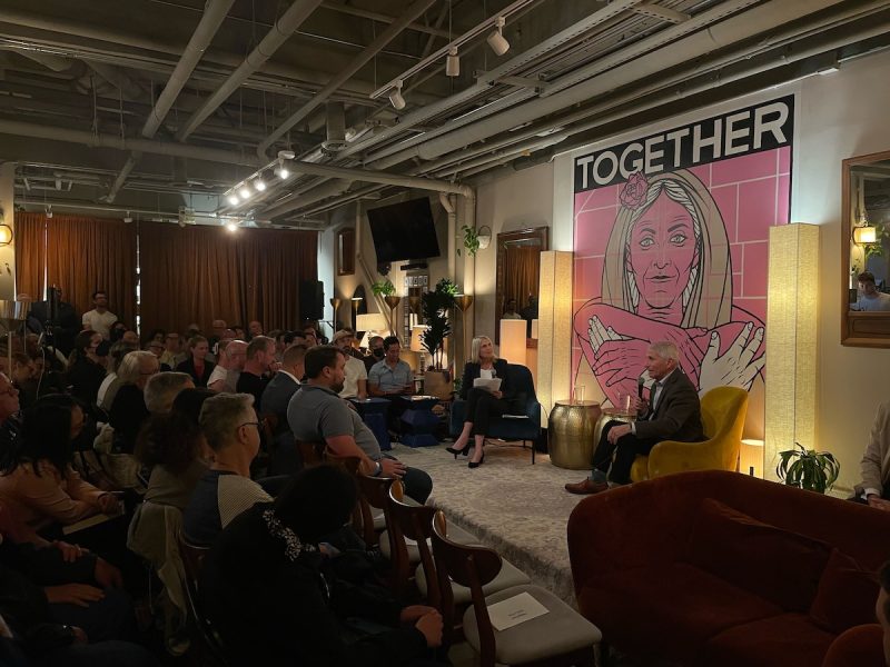 A group of people sit in an indoor venue with low lighting, attentively listening to a speaker. A large artwork with the word "TOGETHER" features prominently in the background.