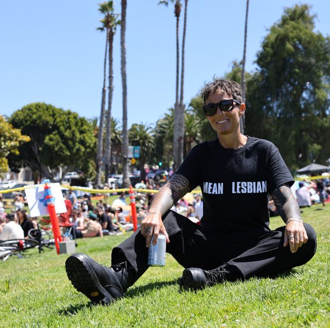 Person wearing sunglasses, a black "MEAN LESBIAN" t-shirt, and black boots, sits on a grassy area holding a drink can, with trees and a crowd in the background.