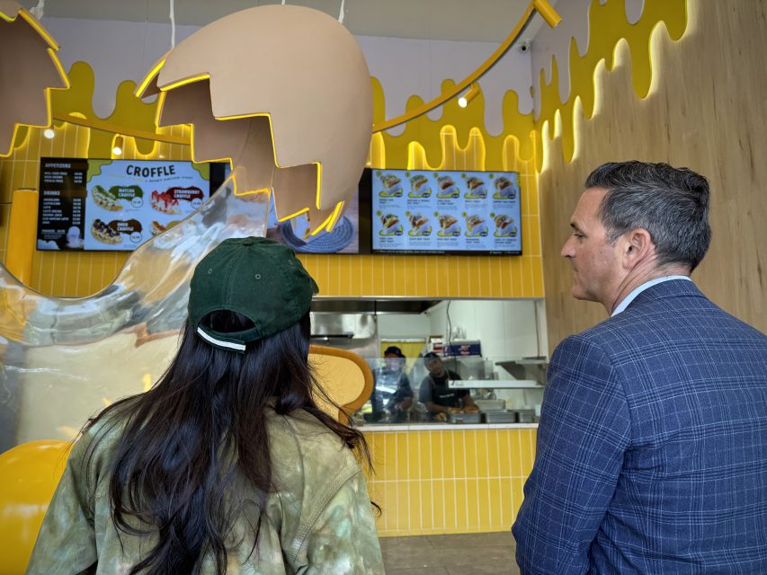 兩個人站在一家以雞蛋爲主題裝飾的咖啡館裡，看着櫃颱上方電子屏幕上顯示的菜單，工作人員正在工作。