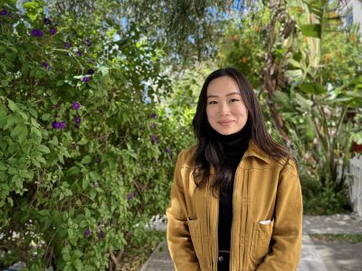 A woman in a yellow jacket stands outdoors, smiling, with a background of lush green foliage and purple flowers.