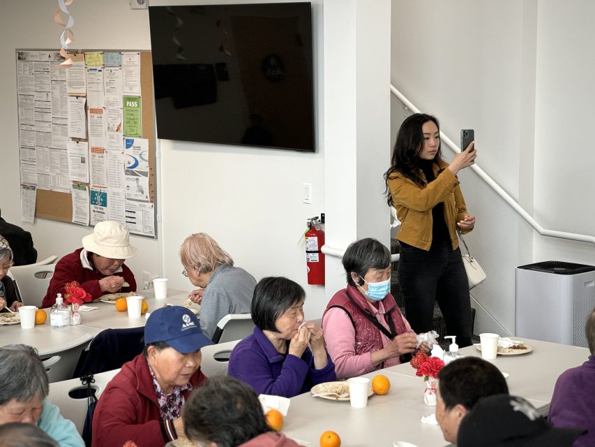 一群老人坐在桌邊用餐。一名女性站在牆邊用手機拍照。背景中可見佈告欄和平闆電視。