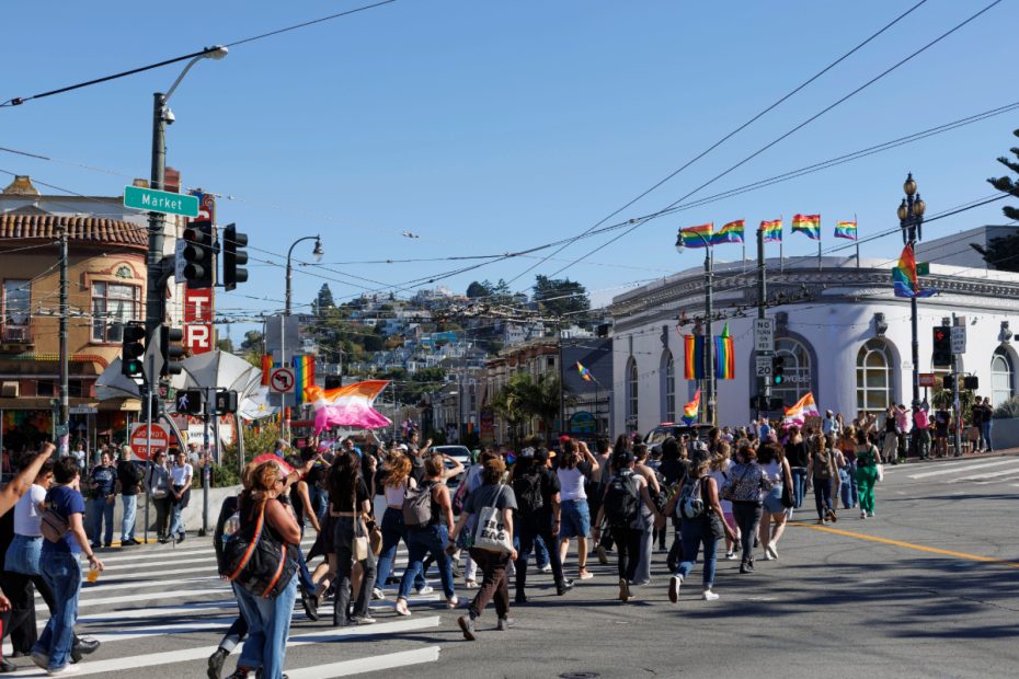 A crowd of people cross a street at a busy intersection on a sunny day, with rainbow flags hanging on buildings and light poles in the background.