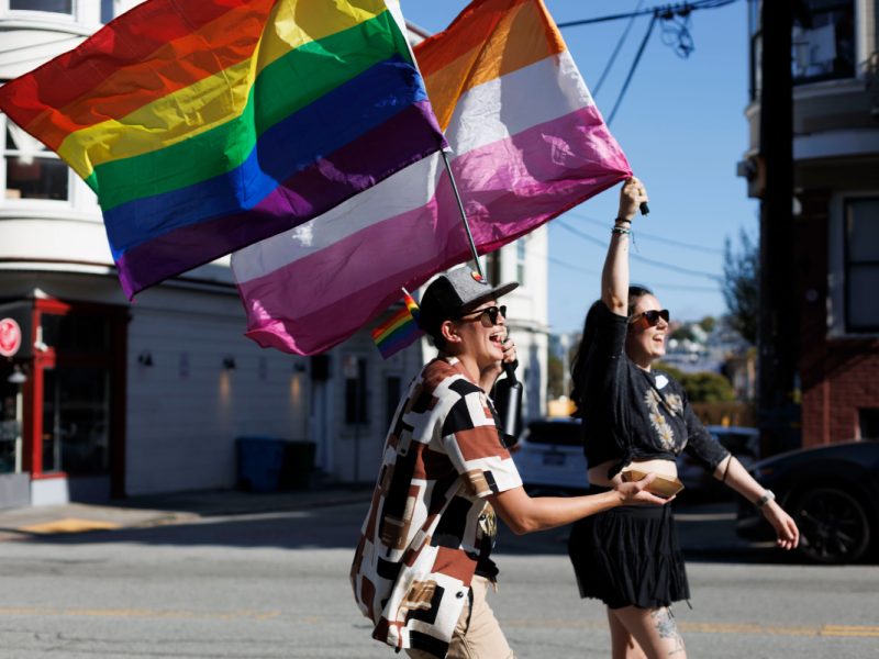 Two individuals joyfully walk down a street carrying a rainbow flag and a lesbian pride flag. One person wears sunglasses and a patterned shirt, the other has a black outfit with shorts.