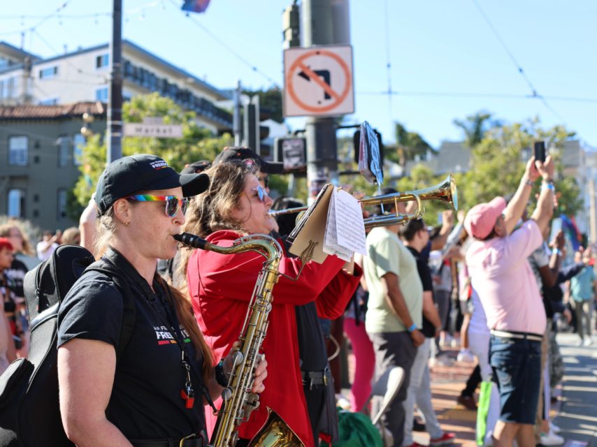 Musicians in a street band, including a saxophonist and trumpeter, perform for a crowd during a sunny day. Some people in the audience are taking photos and recording videos.