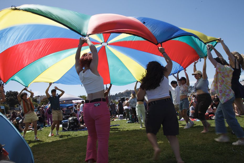 A group of people standing in a circle, lifting a brightly colored parachute in the air at an outdoor gathering on a sunny day.