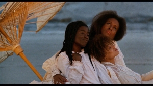 Three people in white clothes sit closely together on a beach under a parasol, looking thoughtfully into the distance.