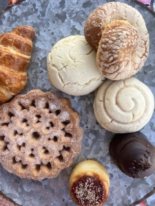 A variety of baked goods arranged on a silver tray, including a croissant, three round breads, a sugar-dusted cookie with a geometric pattern, a jam-filled cookie, and a chocolate-covered confection.
