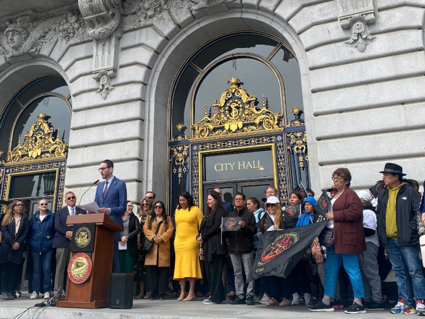 A man speaks at a podium in front of City Hall with a large group of people standing behind and beside him. Some hold a banner, and the ornate building facade is visible.