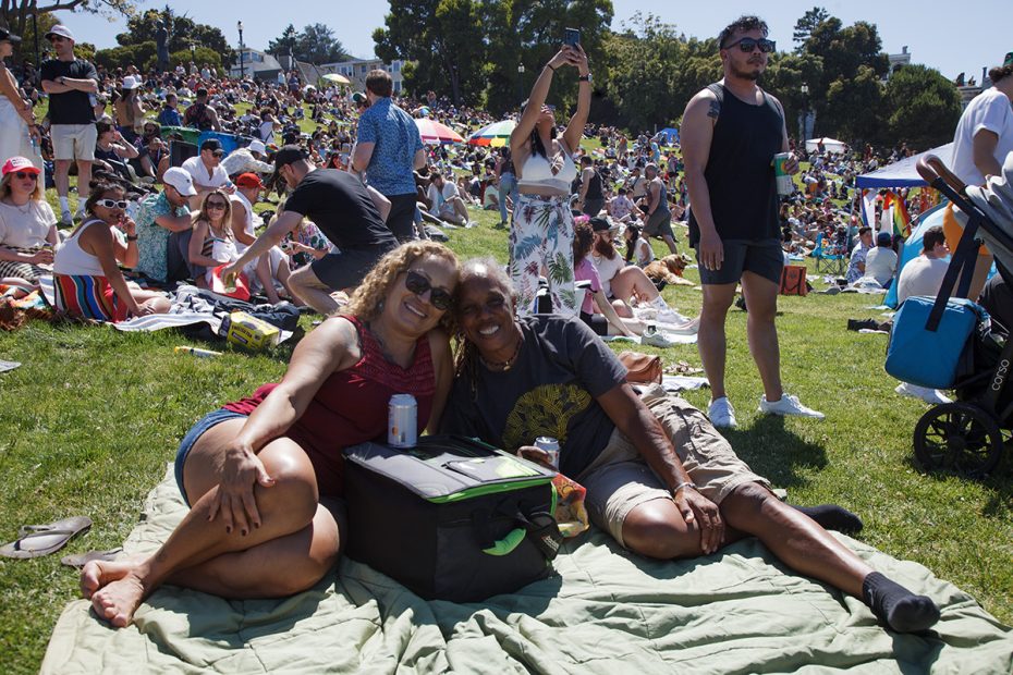 Two people sit on a blanket, smiling at the camera, as a large crowd enjoys a sunny day at an outdoor event. .