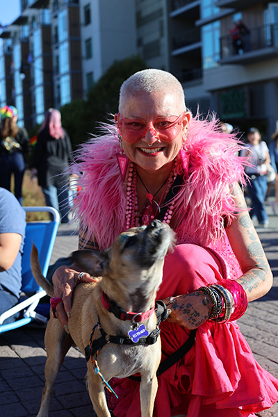 A person in bright pink attire and glasses smiles while kneeling next to a small dog wearing a purple tag, with buildings in the background.