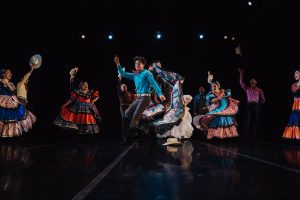 A group of dancers in colorful traditional attire performs energetic movements on a dimly lit stage.