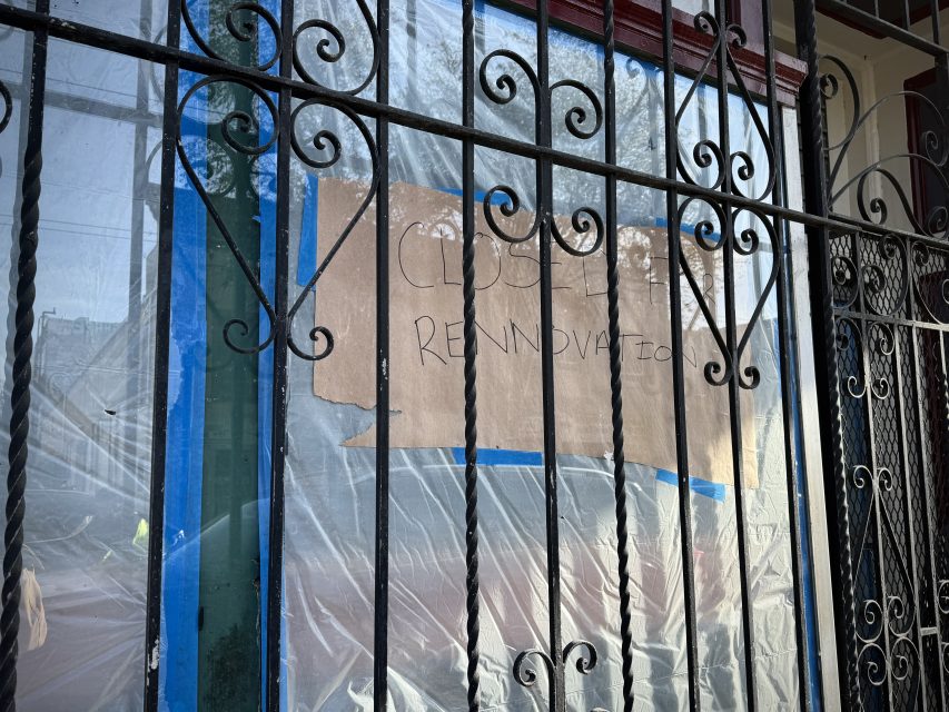 Storefront with a "Closed Renovation" sign made from cardboard and handwritten text, behind an iron gate. The window is covered with plastic sheeting.