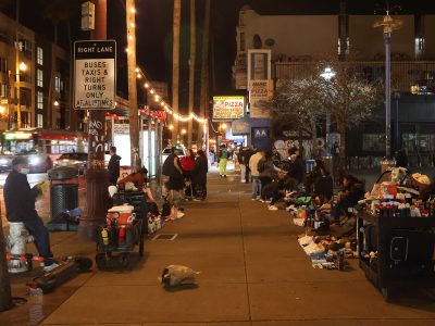 A nighttime street scene with people gathered along the sidewalk selling various items under illuminated string lights. A bus is visible in the background, and storefronts are lit up.