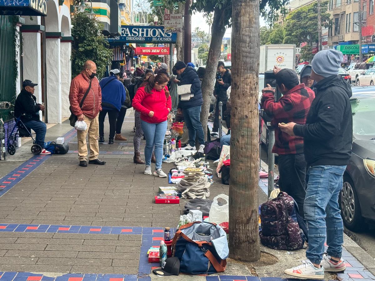 People are browsing and selling various items displayed on the sidewalk of a busy street with shops in the background.