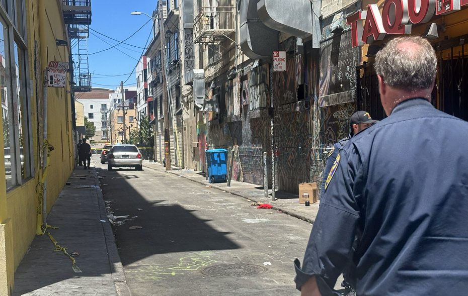 A police officer in uniform stands in an alleyway where yellow caution tape is up. A white car and various graffiti are visible, and two other officers are seen farther down the alley.