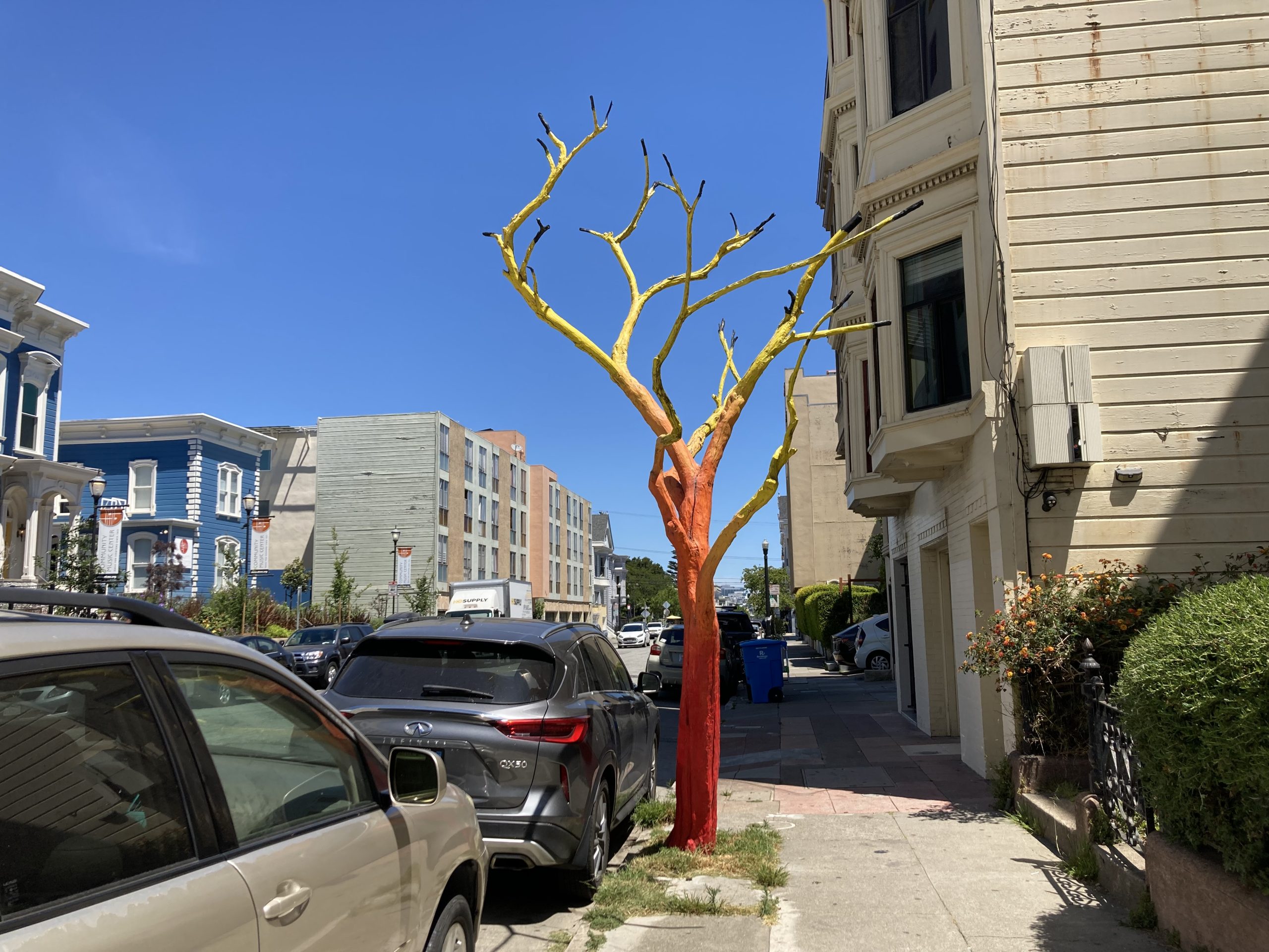 A leafless tree on a city sidewalk is painted with yellow branches and a red trunk. Cars are parked along the street, and buildings are visible in the background.