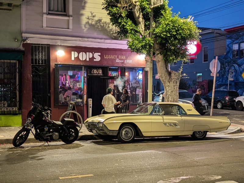 A street scene at night featuring an old-fashioned bar named "Pop's" with a motorcycle and a vintage car parked outside.