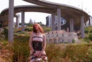 A person with red hair stands in a grassy area wearing a dress, with a large highway overpass and a house in the background.