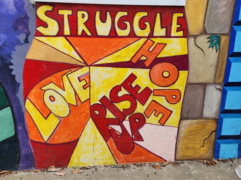 A colorful mural with the words "Struggle," "Hope," "Love," and "Rise Up" in large, bold letters on a textured wall.