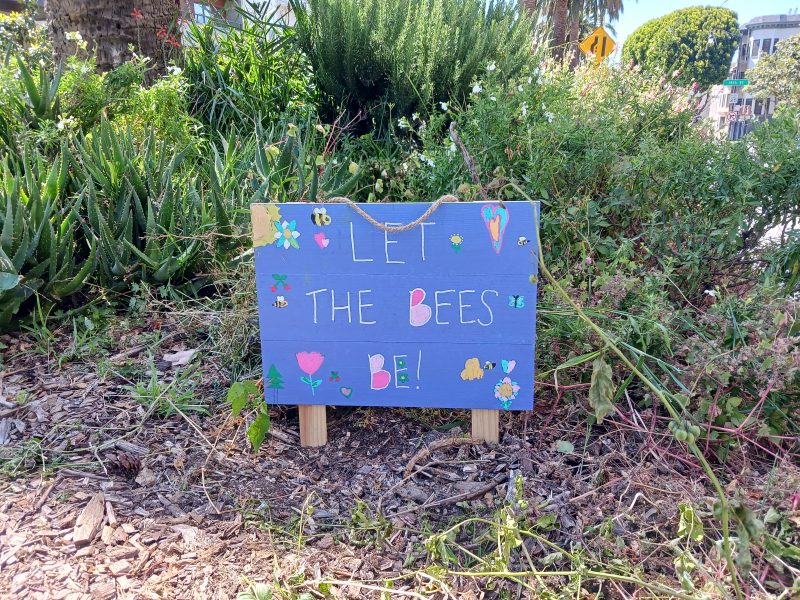 A blue wooden sign with colorful drawings and text that says "Let the bees be!" is placed among greenery and plants in a garden.