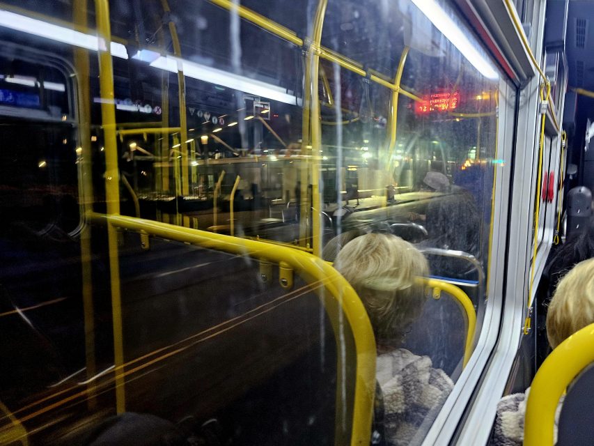 View from inside a bus at night, showing the bus interior, yellow handrails, and a reflection in the window of passengers and the street outside.