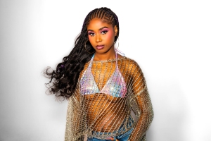 A woman with long braided hair and colorful makeup wears a sparkly bikini top and a mesh overlay shirt against a white background.