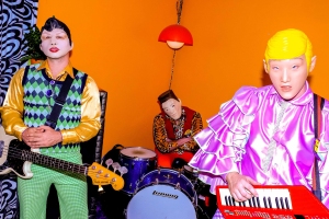 Three musicians in colorful, eccentric outfits perform in a room with bright yellow walls. One plays a bass guitar, another is at a drum set, and the third plays a keyboard.