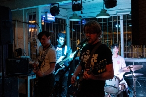 A four-member band performs in a dimly lit venue. Two members play guitars and sing, one plays bass, and one plays drums.