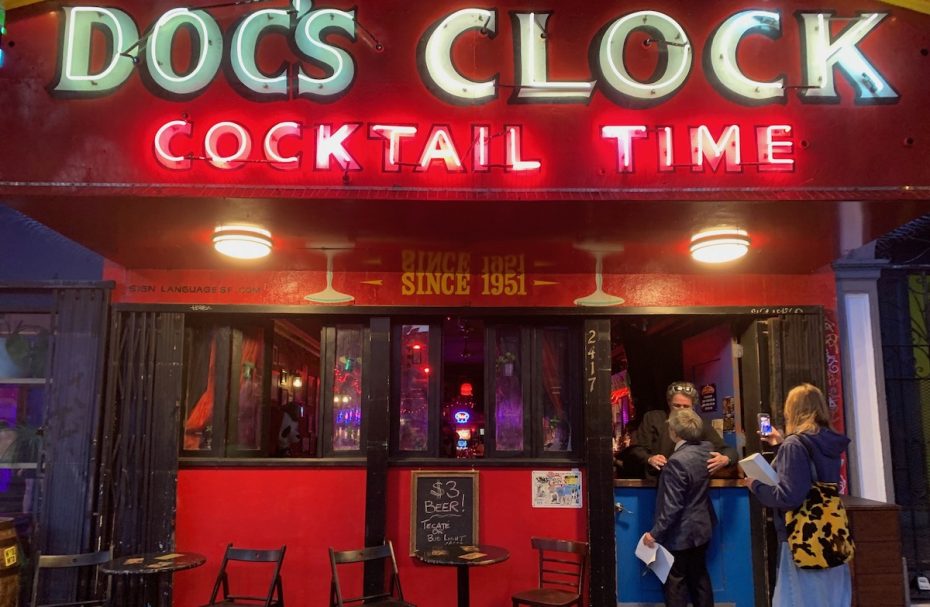 Exterior view of a bar named "Doc's Clock" with bright neon signs reading "Cocktail Time." A few people stand at the bar's entrance, and a chalkboard advertises a $3 beer special.