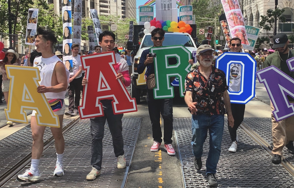 A group of people holding large cardboard letters spelling "AARON" walks down a street during a parade, with a truck decorated with balloons behind them.